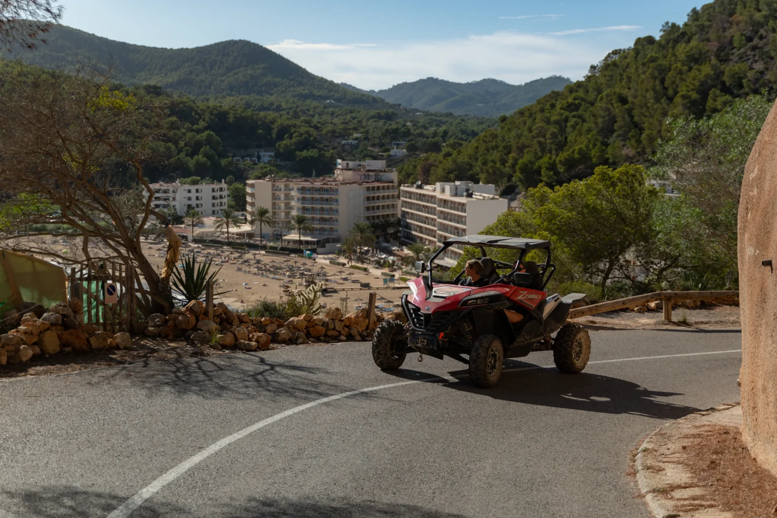 Off-road voertuig dat een bocht maakt op een kronkelige weg langs groene heuvels en een baai met strandligstoelen.