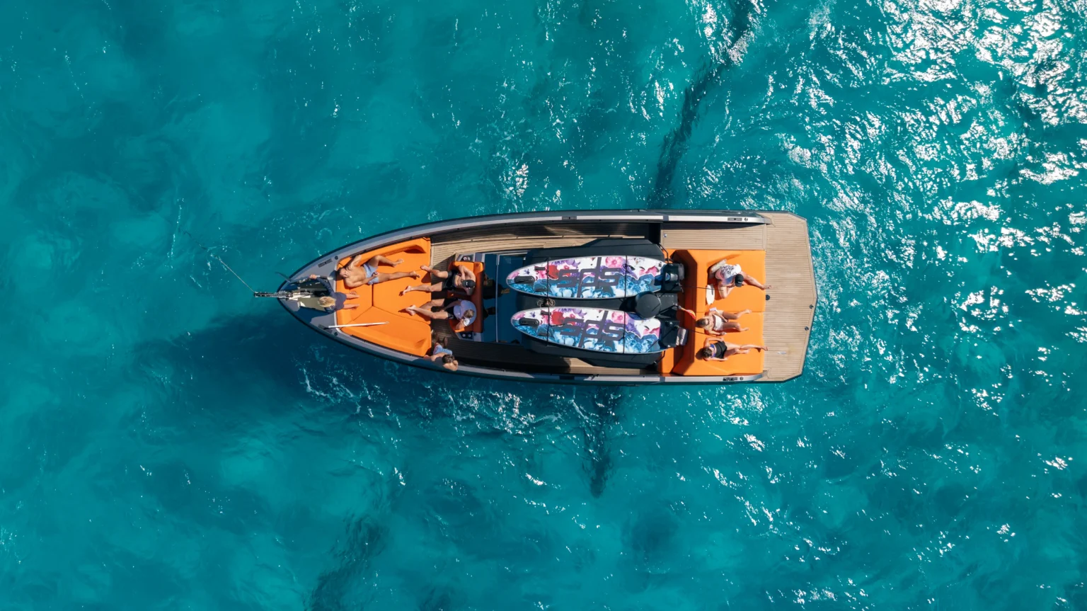 Luxe boot met oranje kussens en twee kleurrijke surfboards op helder turquoise water gezien vanaf boven