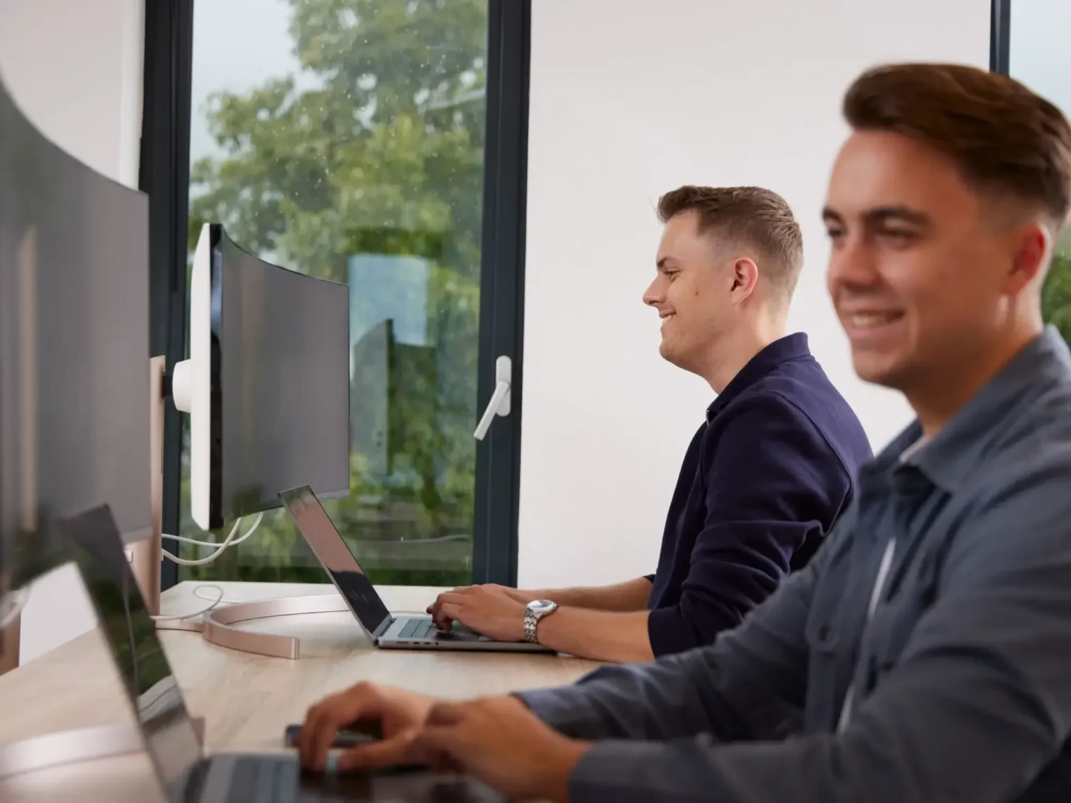 Twee jonge mannen werken geconcentreerd op laptops aan een lange houten tafel in een lichte kantoorruimte met grote ramen.