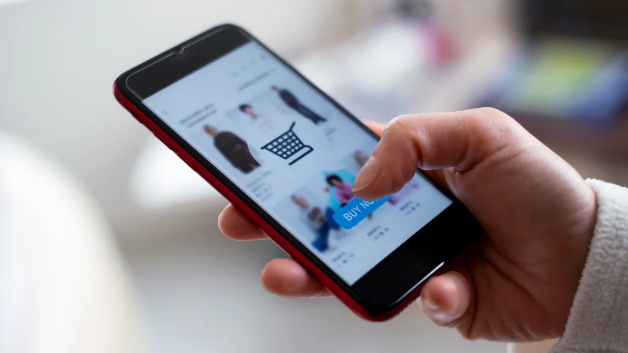 Hand houdt smartphone met online winkelweergave, producten in raster en knop 'BUY NOW'.