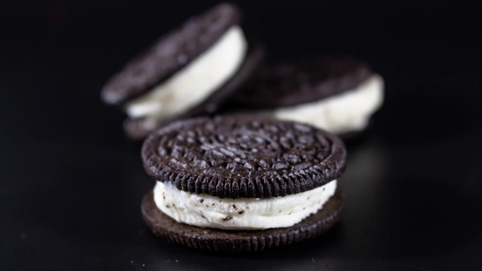 Enkele donkerbruine chocoladekoekjes met witte romige vulling en gedetailleerd patroon op een donkere achtergrond