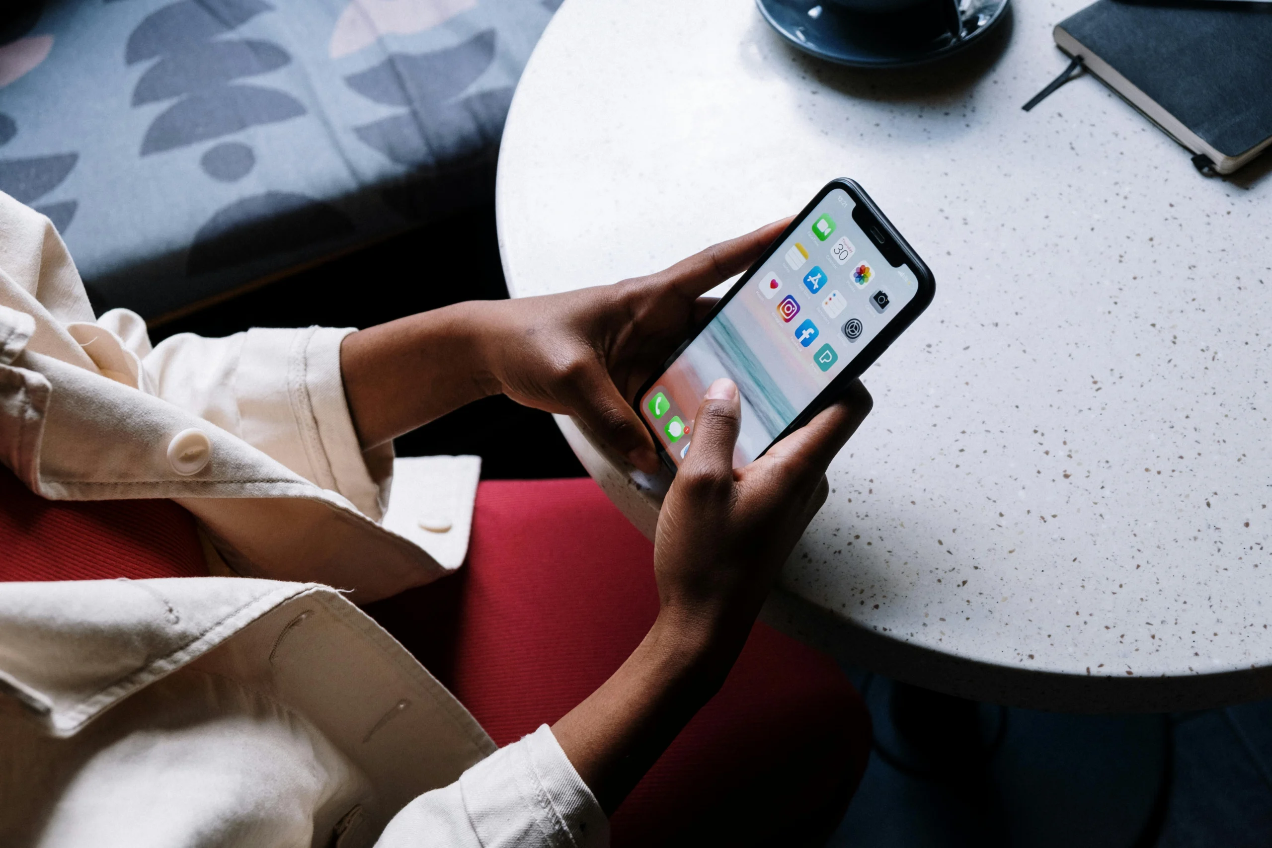 Persoon met lichte jas en roze broek houdt smartphone vast aan ronde tafel met notitieboekje en kopje